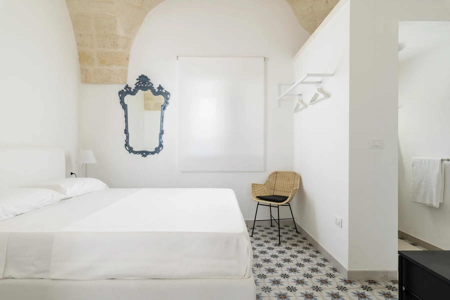 Un dormitorio luminoso con ropa de cama blanca, un espejo en la pared y suelo de baldosas estampadas.