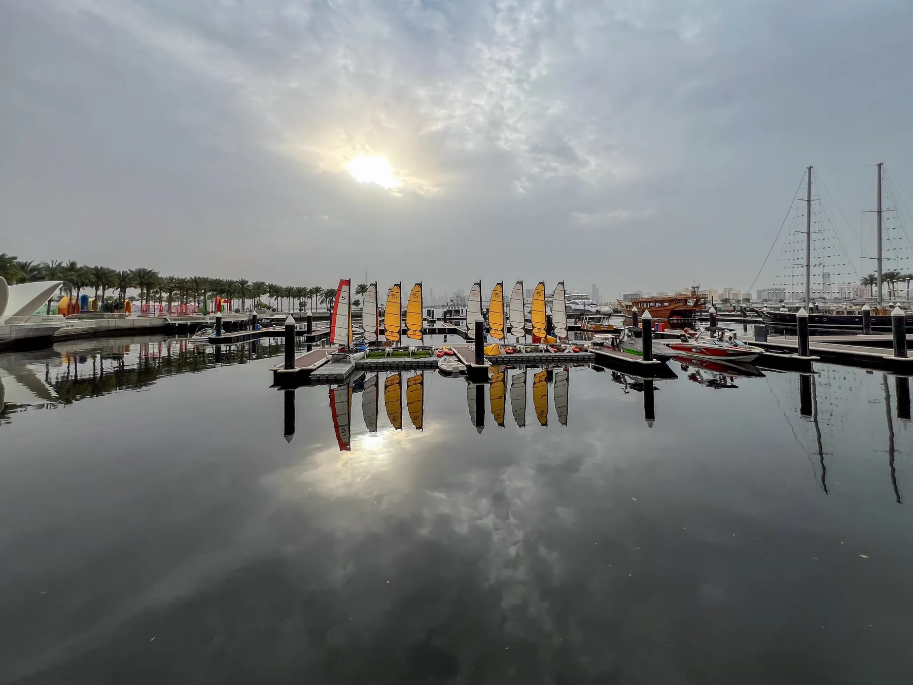 Una marina con veleros de colores reflejados en aguas tranquilas bajo un cielo nublado.