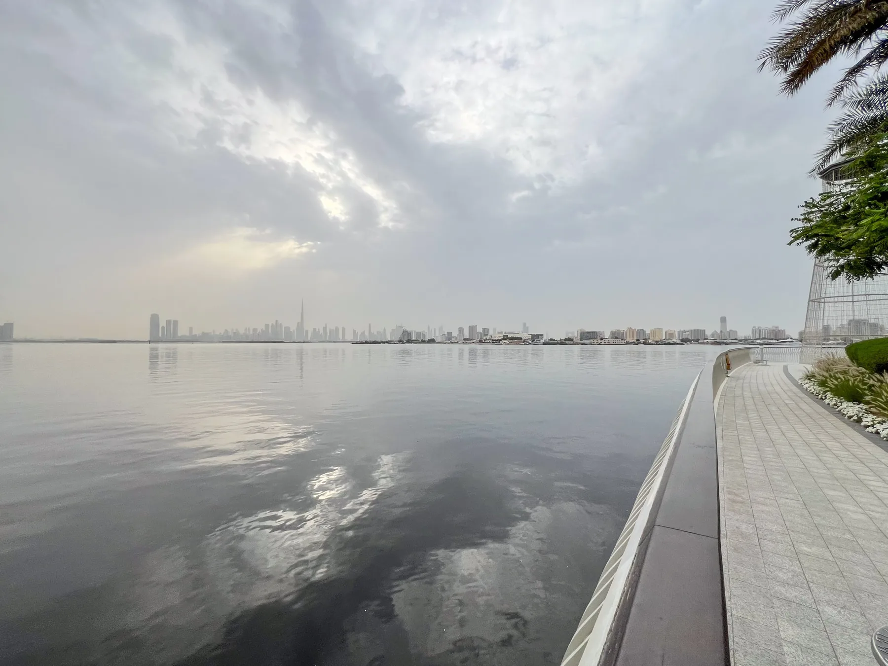 Un paseo junto al agua tranquila se curva frente a un horizonte urbano distante.