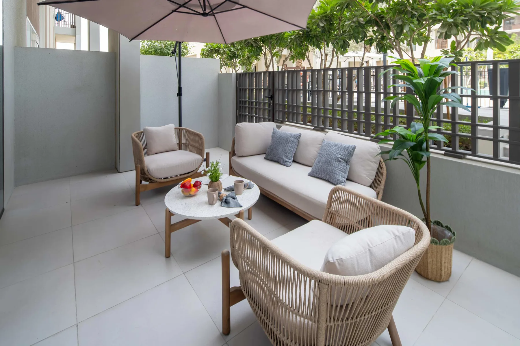 Una zona de estar al aire libre con suelo de baldosas, sillones con cojines, una mesa y una sombrilla.