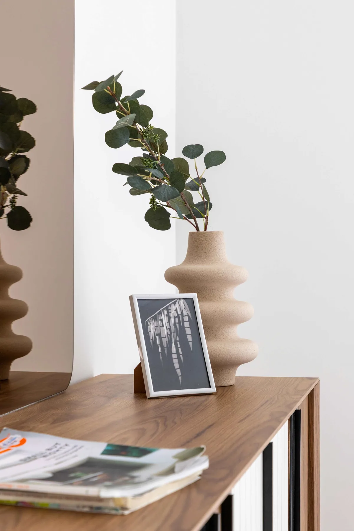 Una mesa de madera muestra una lámina enmarcada, un jarrón con ramas frondosas y revistas.