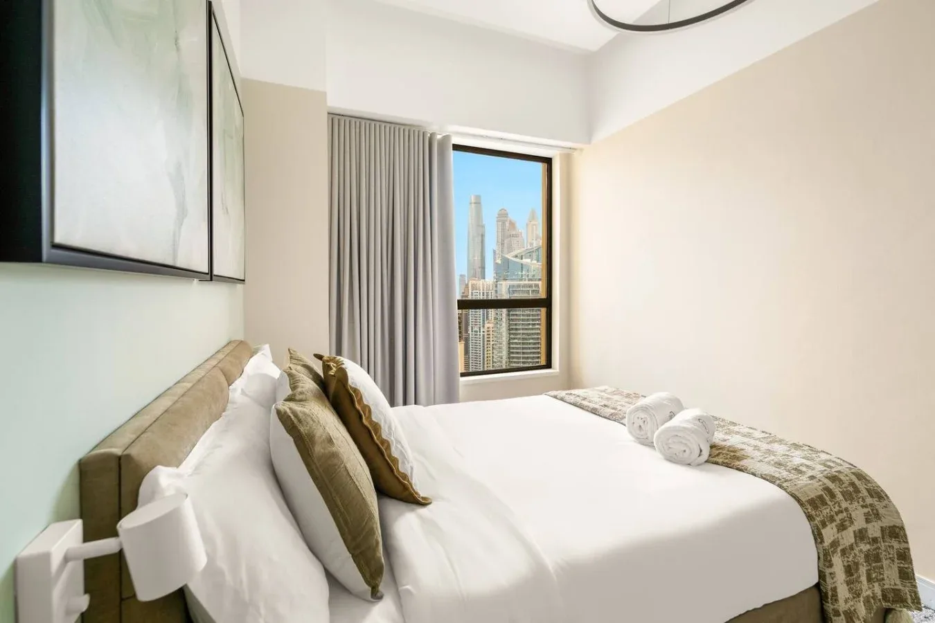 A bedroom with a neatly made bed and a window overlooking city buildings.