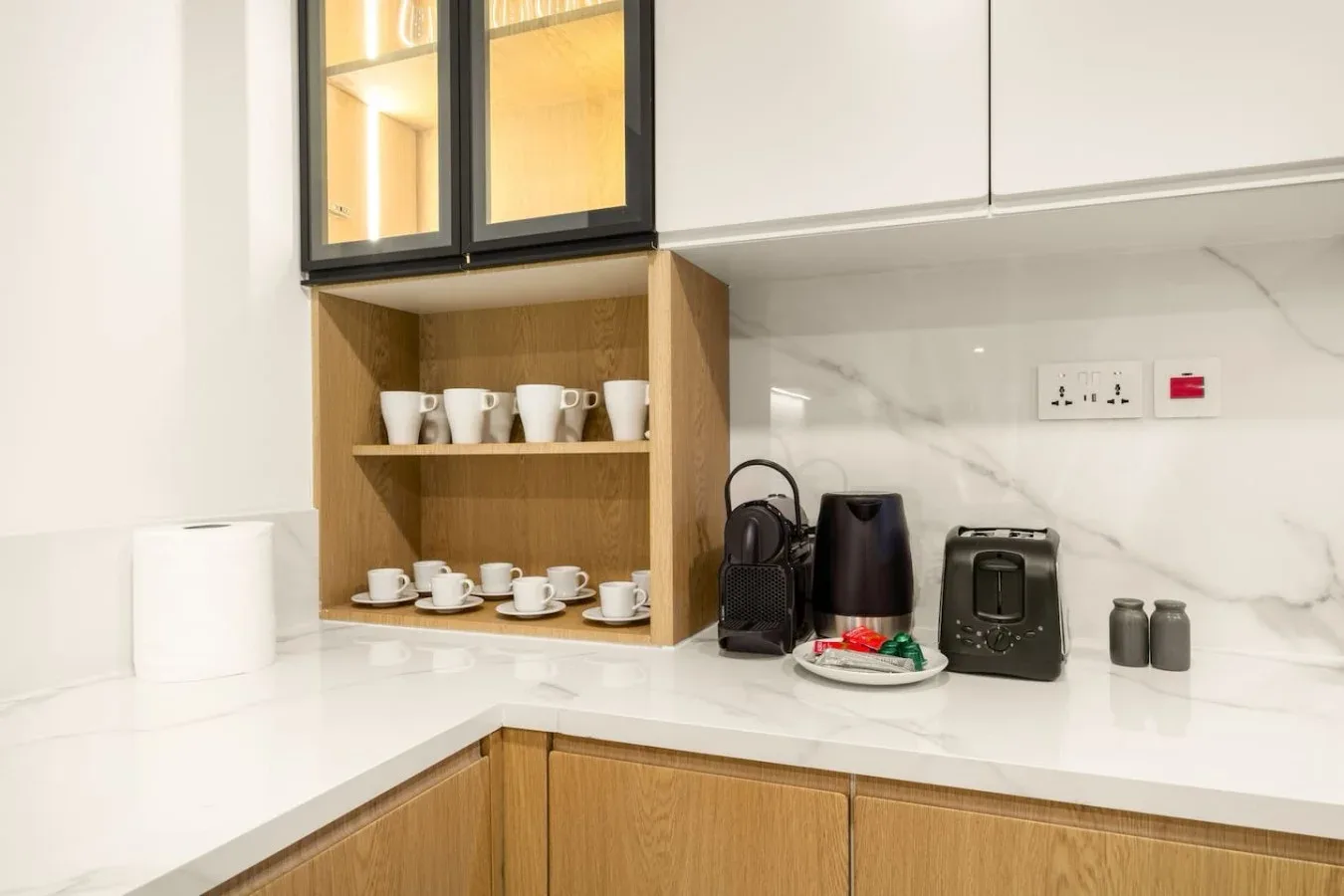 A modern kitchenette counter displays cups, a kettle, and a coffee maker.