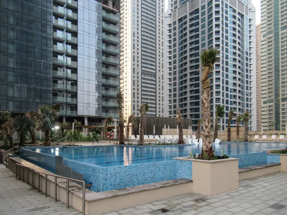 An outdoor swimming pool sits among tall residential buildings and palm trees.