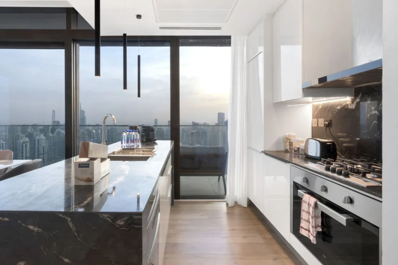 A modern kitchen with a dark island and floor-to-ceiling windows overlooking the city.