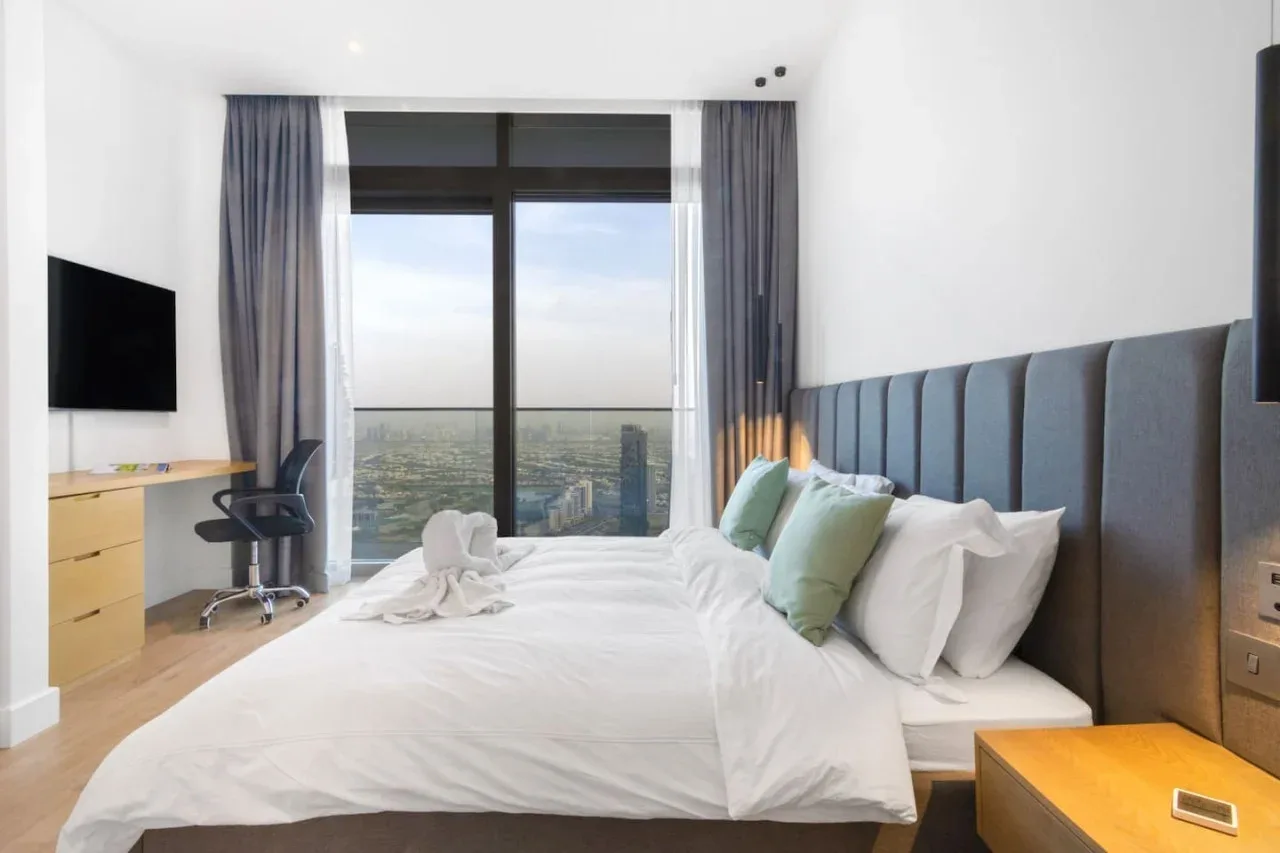 A bedroom with a neatly made bed, gray curtains, and a large window overlooking the city.