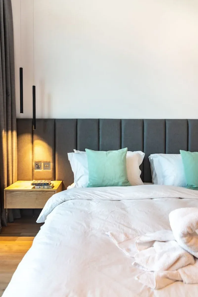 A bedroom with white bedding, aqua pillows, and a dark upholstered headboard.