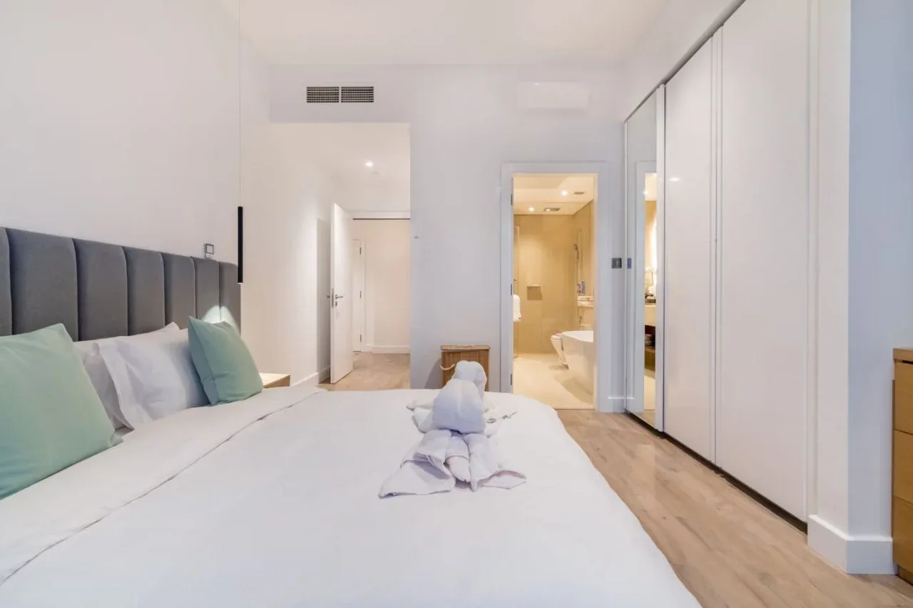A bright bedroom with a neatly made bed and an open doorway to the bathroom.