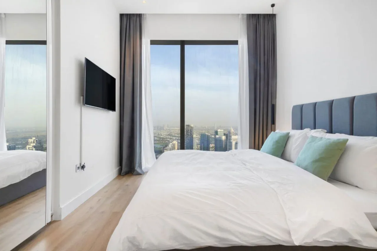 A bedroom with a large bed, a wall-mounted television, and a city view through the window.