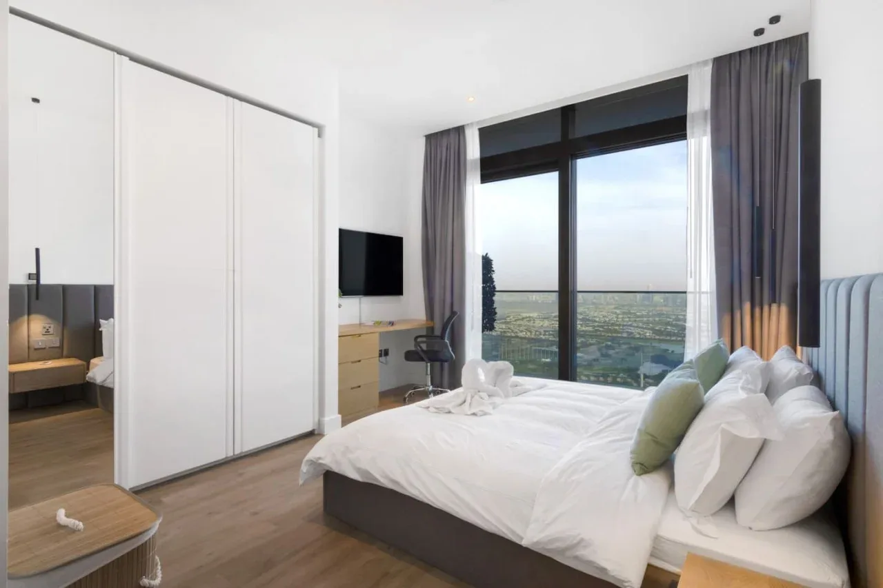 A bright bedroom with a bed beside a large window overlooking the city.