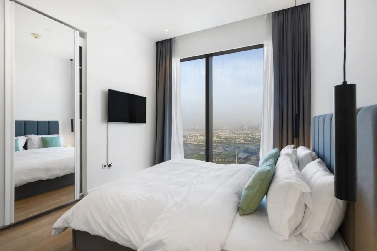 A bright bedroom with a bed beside a large window overlooking the city.