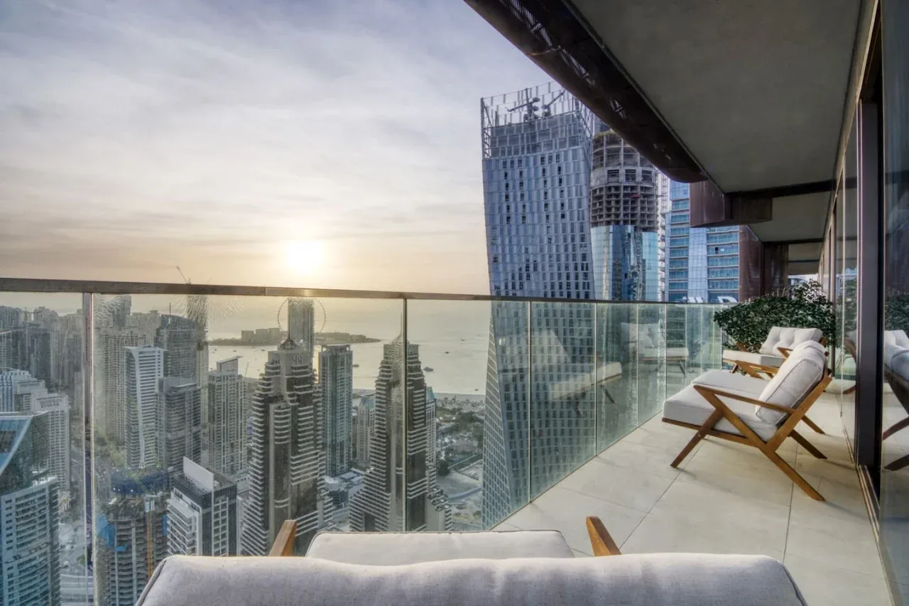 A balcony seating area overlooks high-rise buildings and the water at sunset.