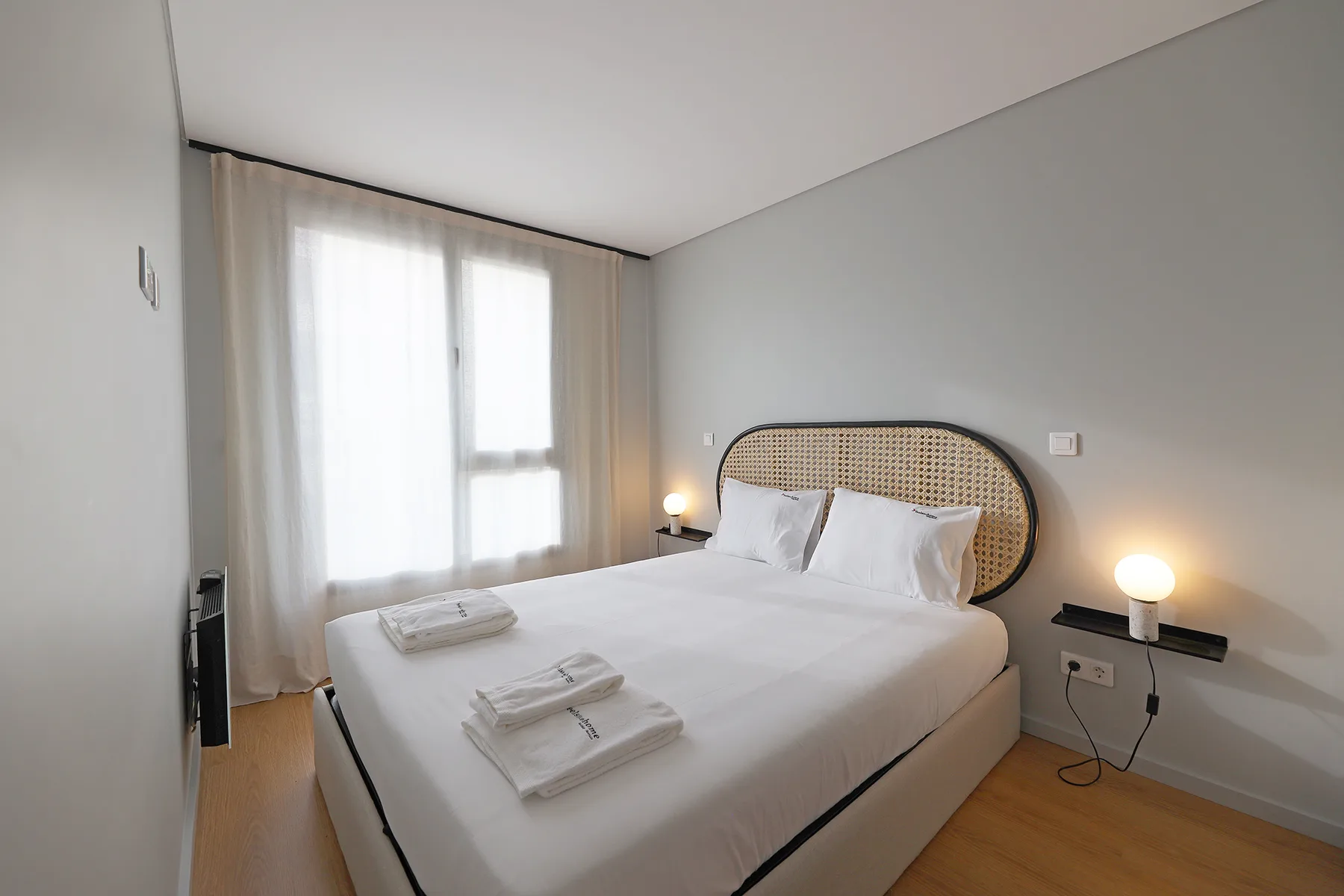 A bedroom with a double bed, folded towels, and a bright curtained window.