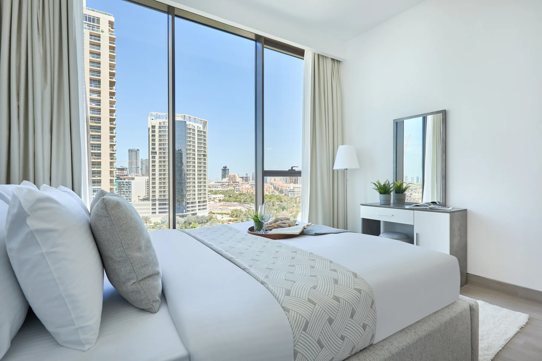 Un dormitorio luminoso con una cama tendida junto a grandes ventanas con vistas a la ciudad.