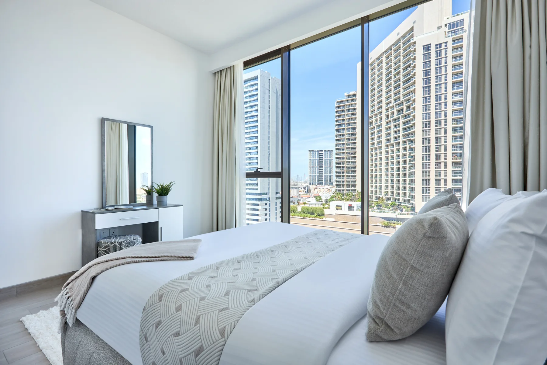 Un dormitorio luminoso con una cama tendida junto a grandes ventanas con vistas a edificios altos.