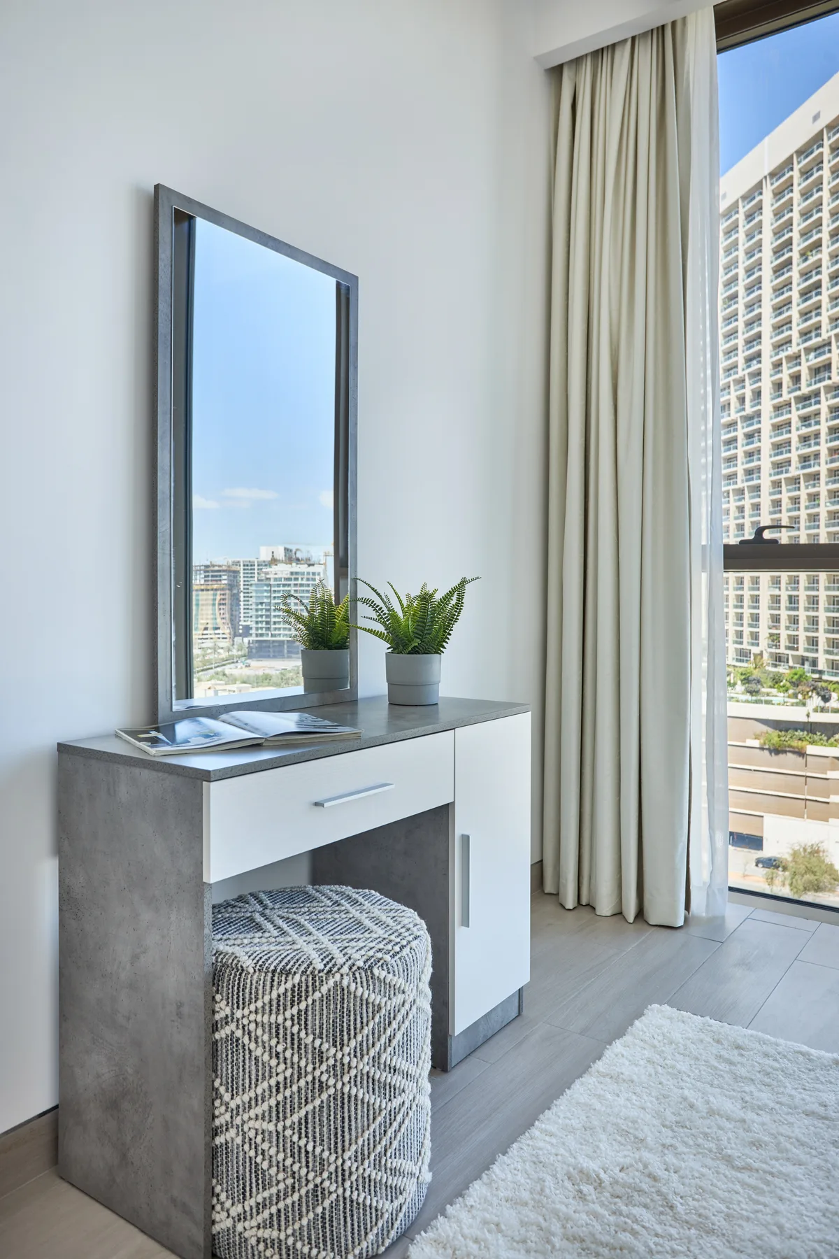 Una esquina luminosa de dormitorio con tocador, asiento estampado y una ventana grande.