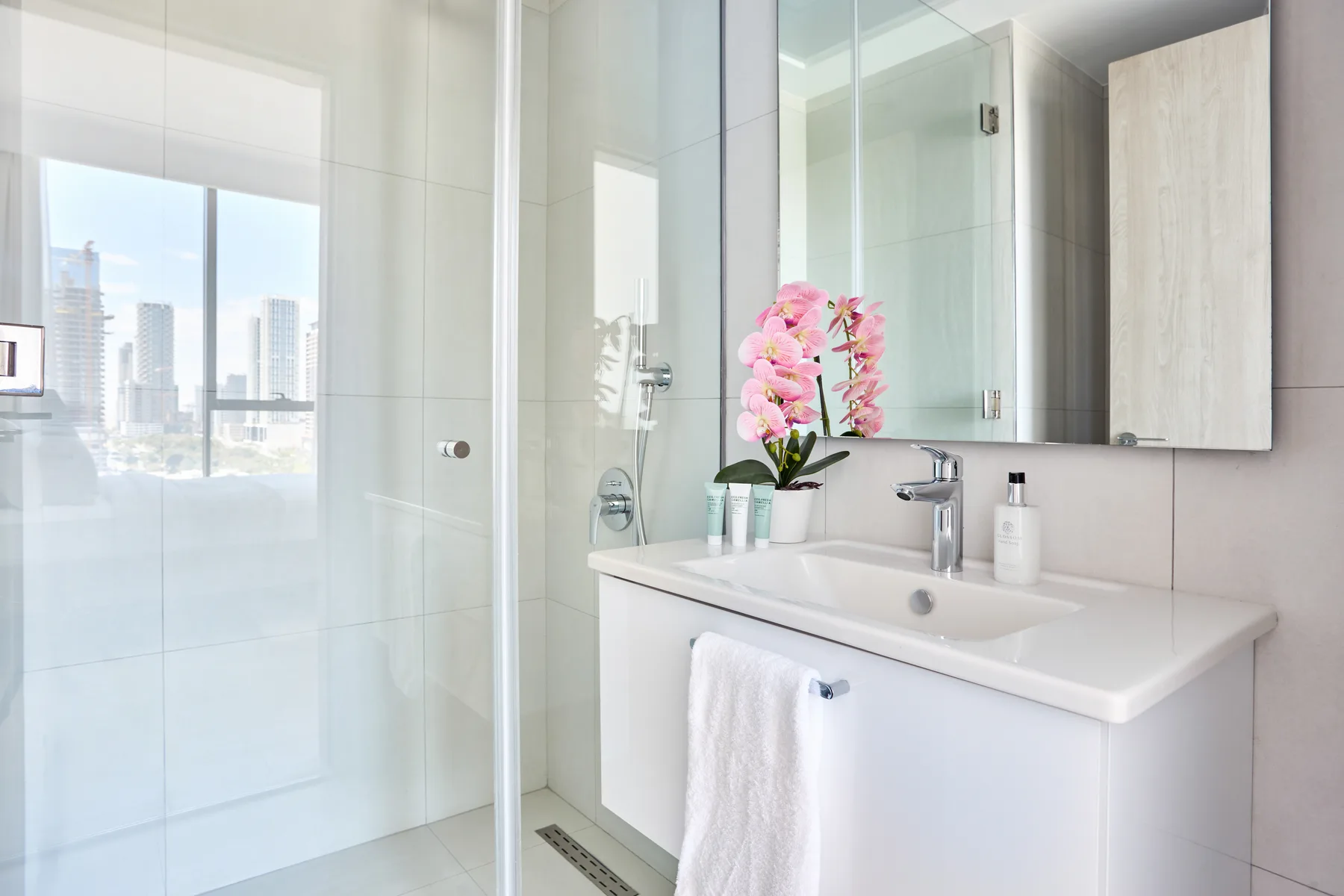 Un baño luminoso tiene una ducha de vidrio y un tocador blanco con flores rosas.