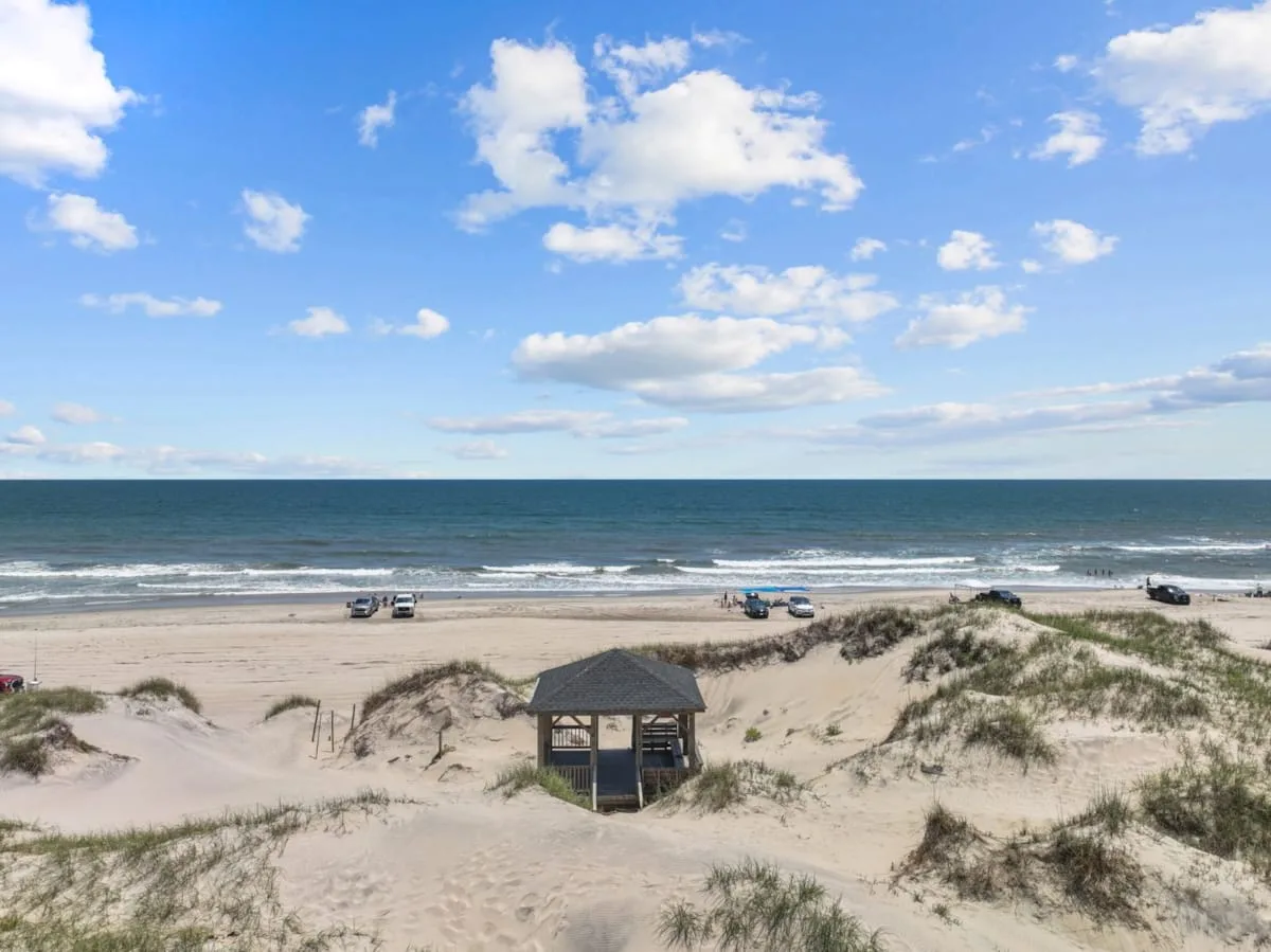 A sandy beach with grassy dunes, ocean waves, and a small wooden shelter.