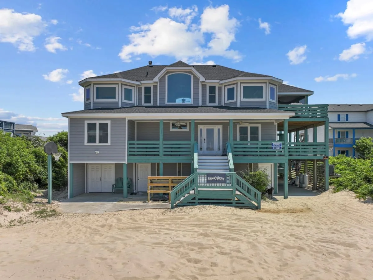 A large gray two-story house with teal porches stands beside a sandy yard.