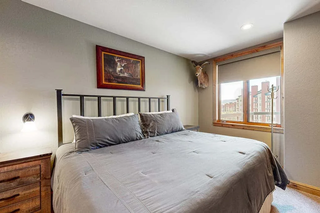 A bedroom with a large bed, framed artwork, and a window overlooking nearby buildings.