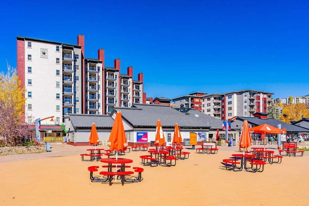 An outdoor seating area with red picnic tables and orange umbrellas in front of apartment buildings.