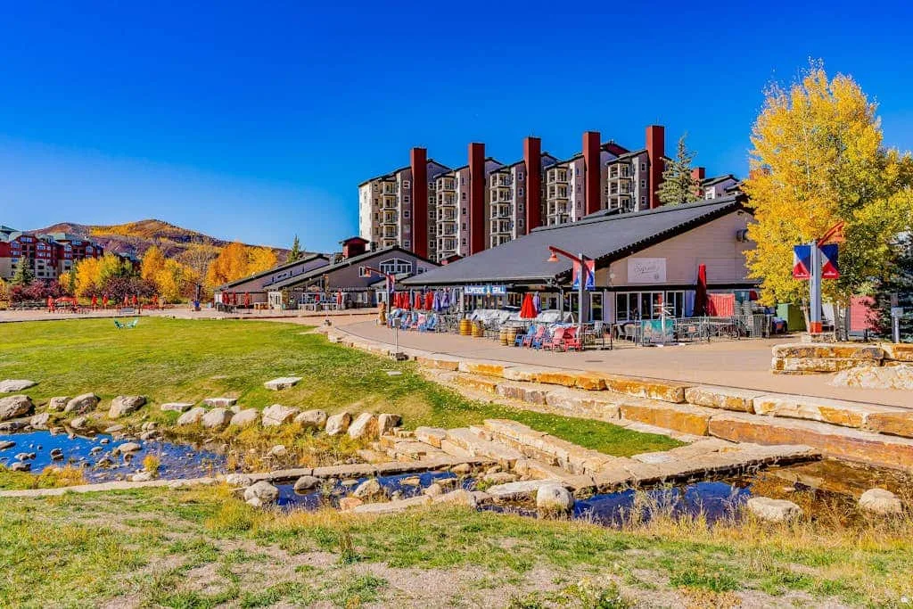 A mountain resort complex stands beside a grassy stream under a clear blue sky.