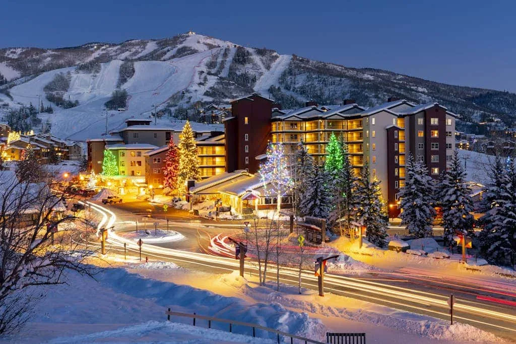 Snow-covered mountain resort buildings glow beside a winding road at dusk.