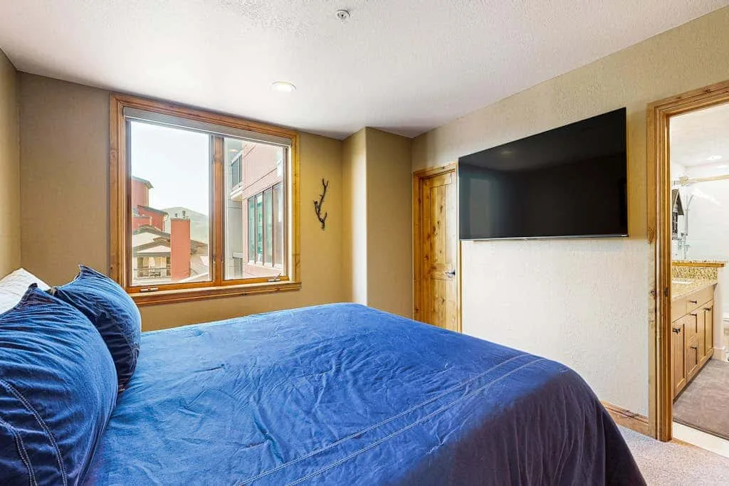 A bedroom with a blue bedspread, a wall-mounted television, and windows.