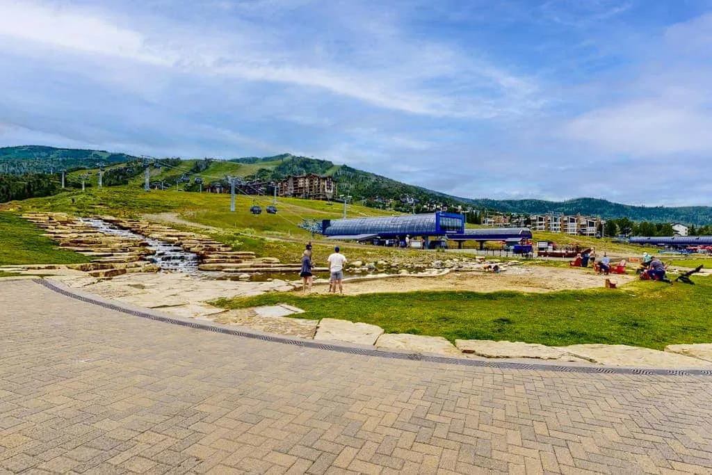 A paved plaza borders a grassy hillside with a long building and distant hills.