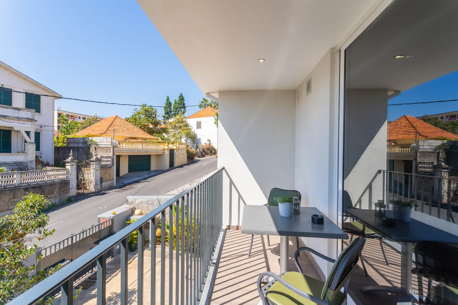 A balcony overlooks a residential street, with a small table and seating beside the glass doors.