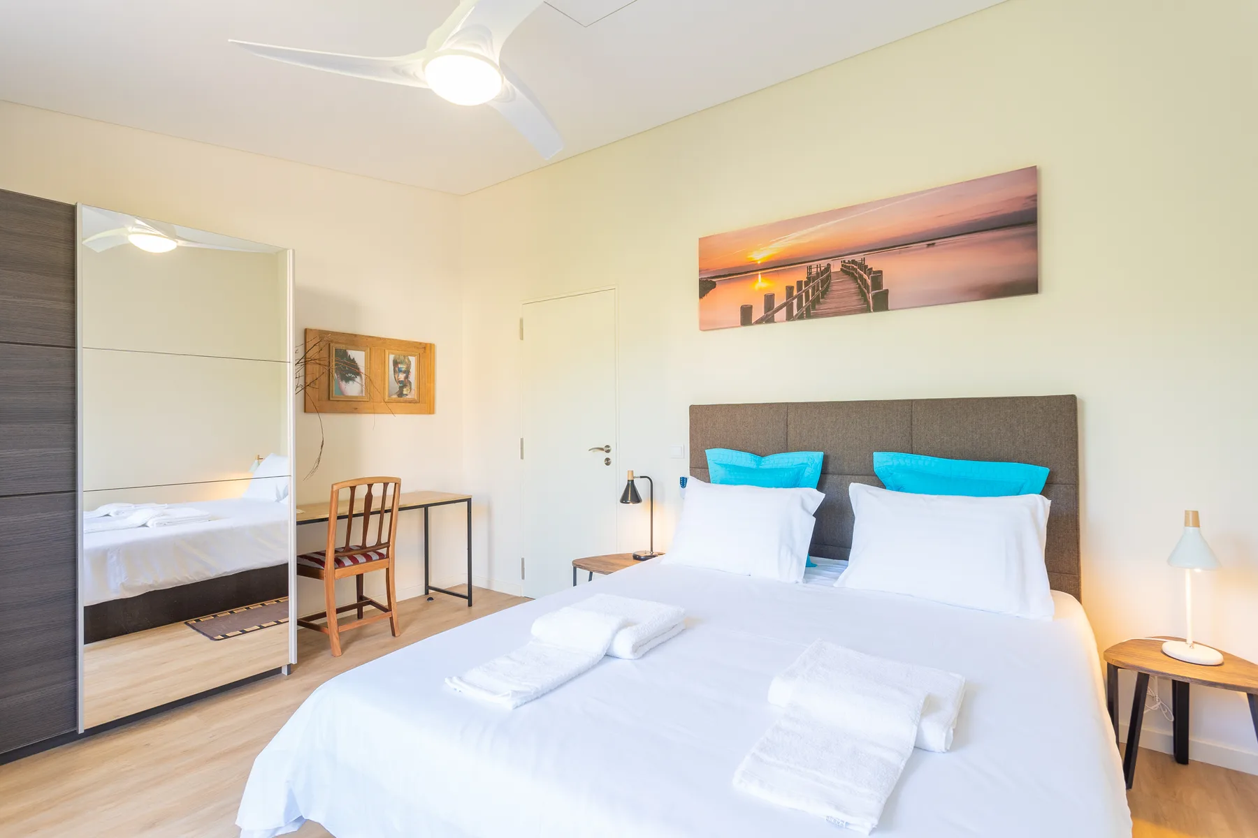 Un dormitorio luminoso con una cama tendida y un escritorio junto al espejo.