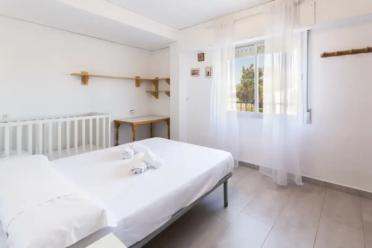 A bright bedroom with a bed beside a window with sheer curtains.