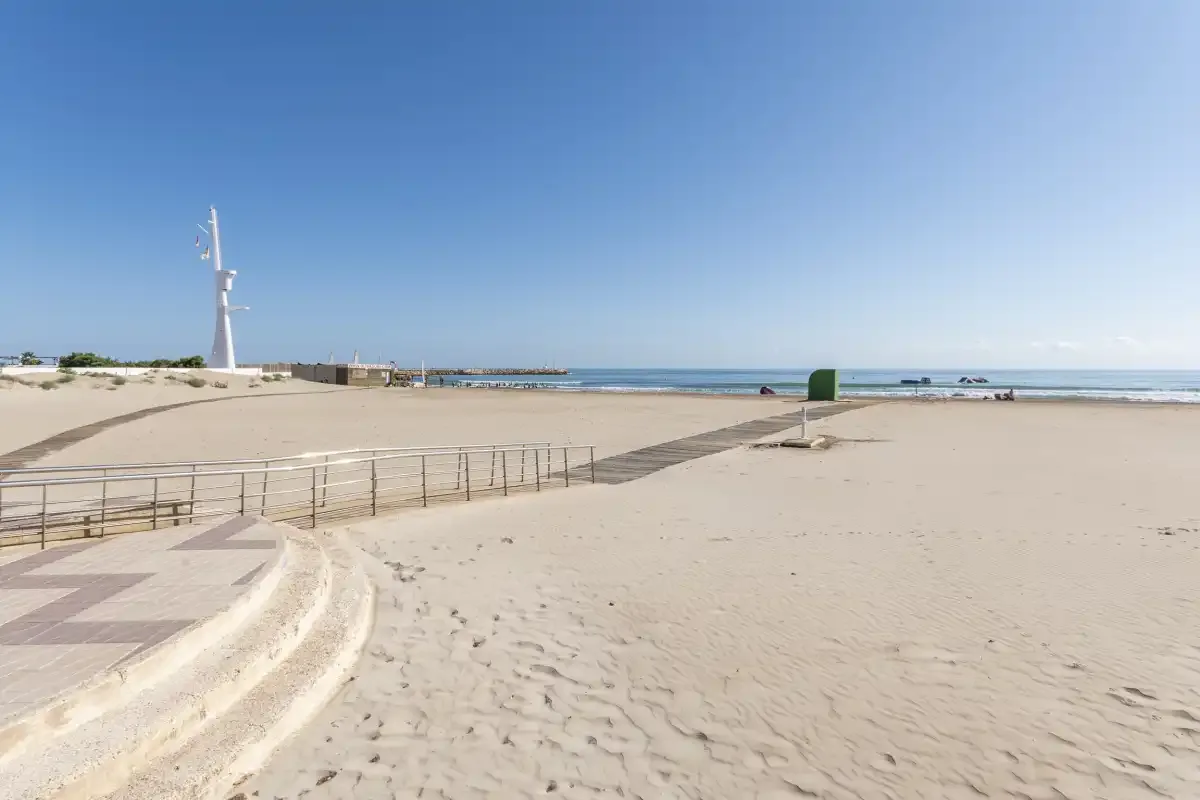 A sandy beach with a paved walkway, railing, and the sea in the distance.