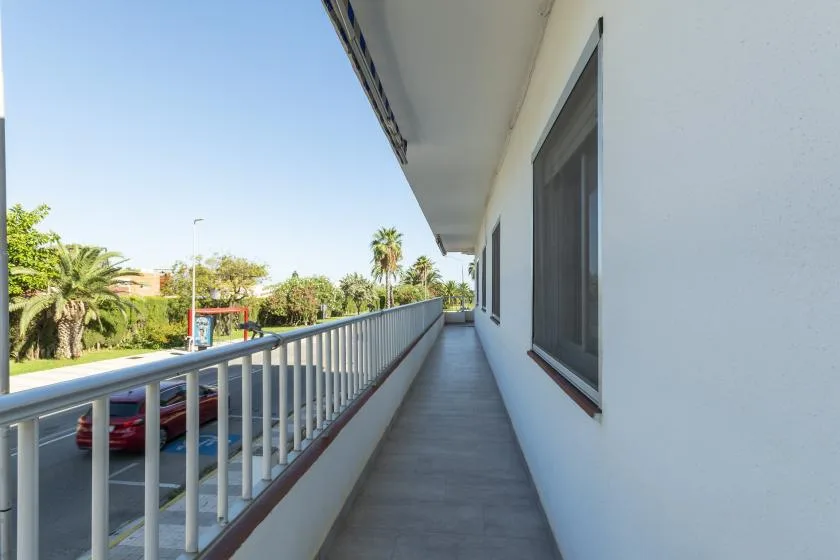 A long outdoor balcony walkway runs alongside a white building, with trees visible beyond the railing.
