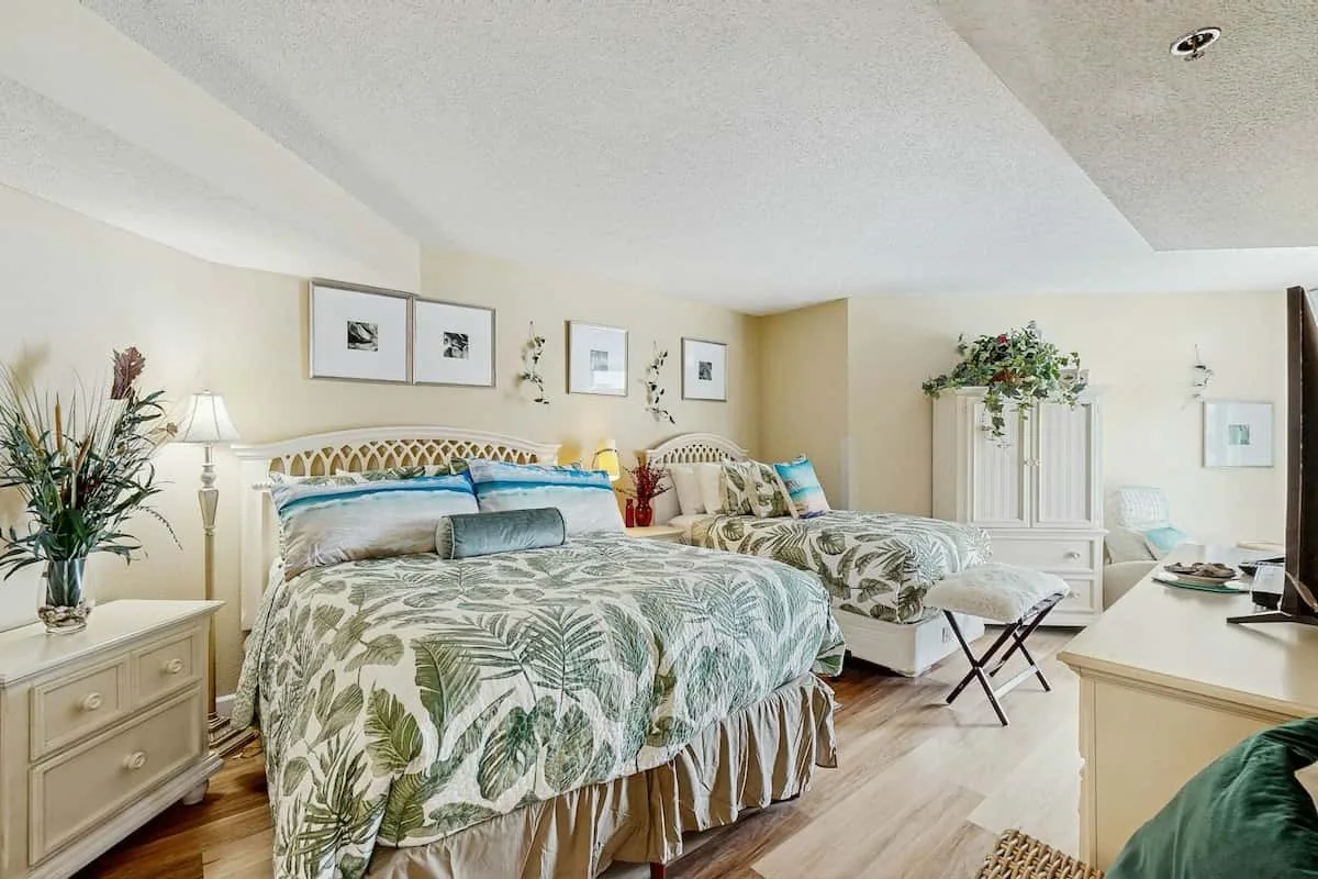 A bright bedroom with two beds, framed pictures, and a dresser.