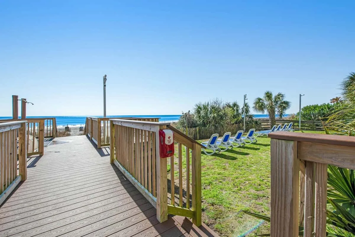 A wooden boardwalk opens onto a grassy beachfront area with lounge chairs and ocean views.