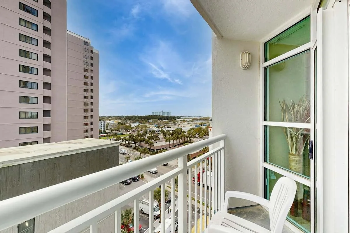 A balcony overlooks nearby buildings and a waterfront under a blue sky.
