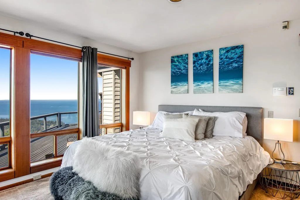 A bright bedroom with a neatly made bed beside large windows overlooking the ocean.