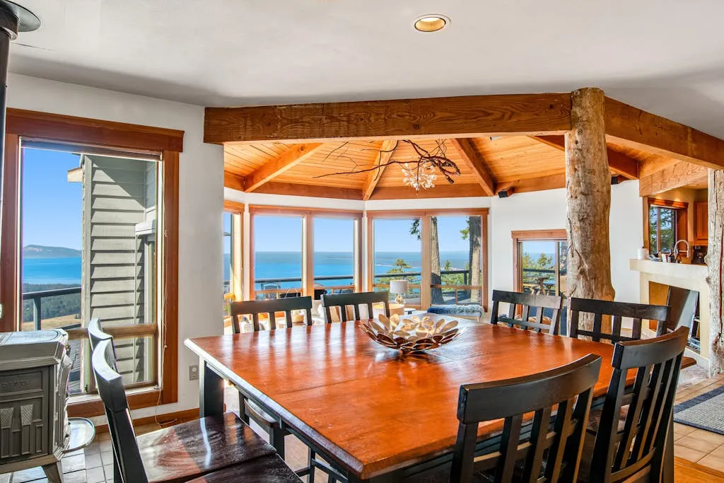 A dining area with a large wooden table and windows overlooking the ocean.