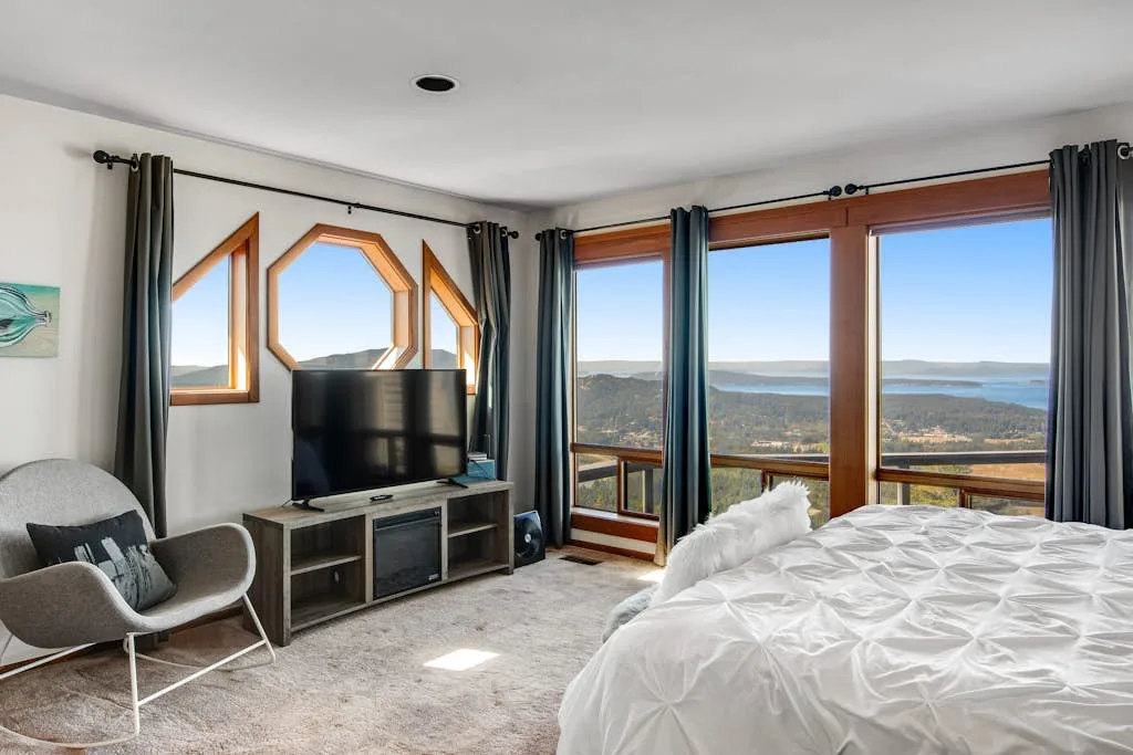 A bedroom with a bed, television, and large windows overlooking a lake.