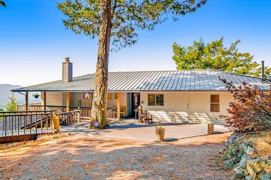 A single-story house with a metal roof stands among trees beside a gravel driveway.