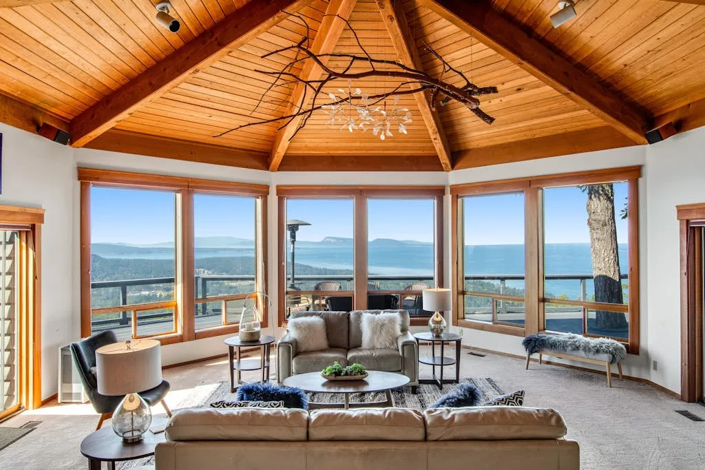 A bright living room with a high wooden ceiling and large windows overlooking the water.