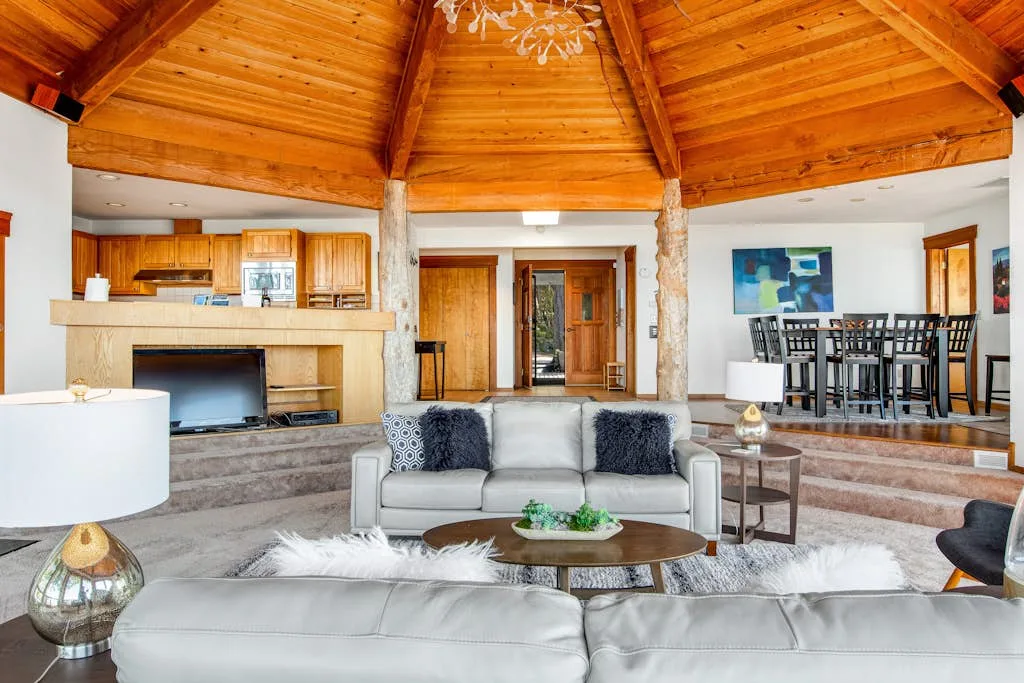 A spacious living room with a vaulted wooden ceiling, seating, and an open kitchen.