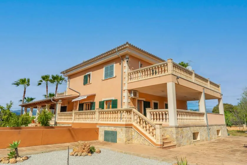 A two-story house with balconies and a gravel driveway under a clear sky.