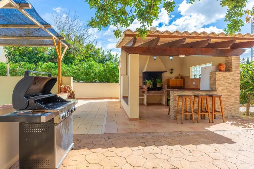 An outdoor kitchen and bar sit beneath a wooden pergola beside a barbecue grill.