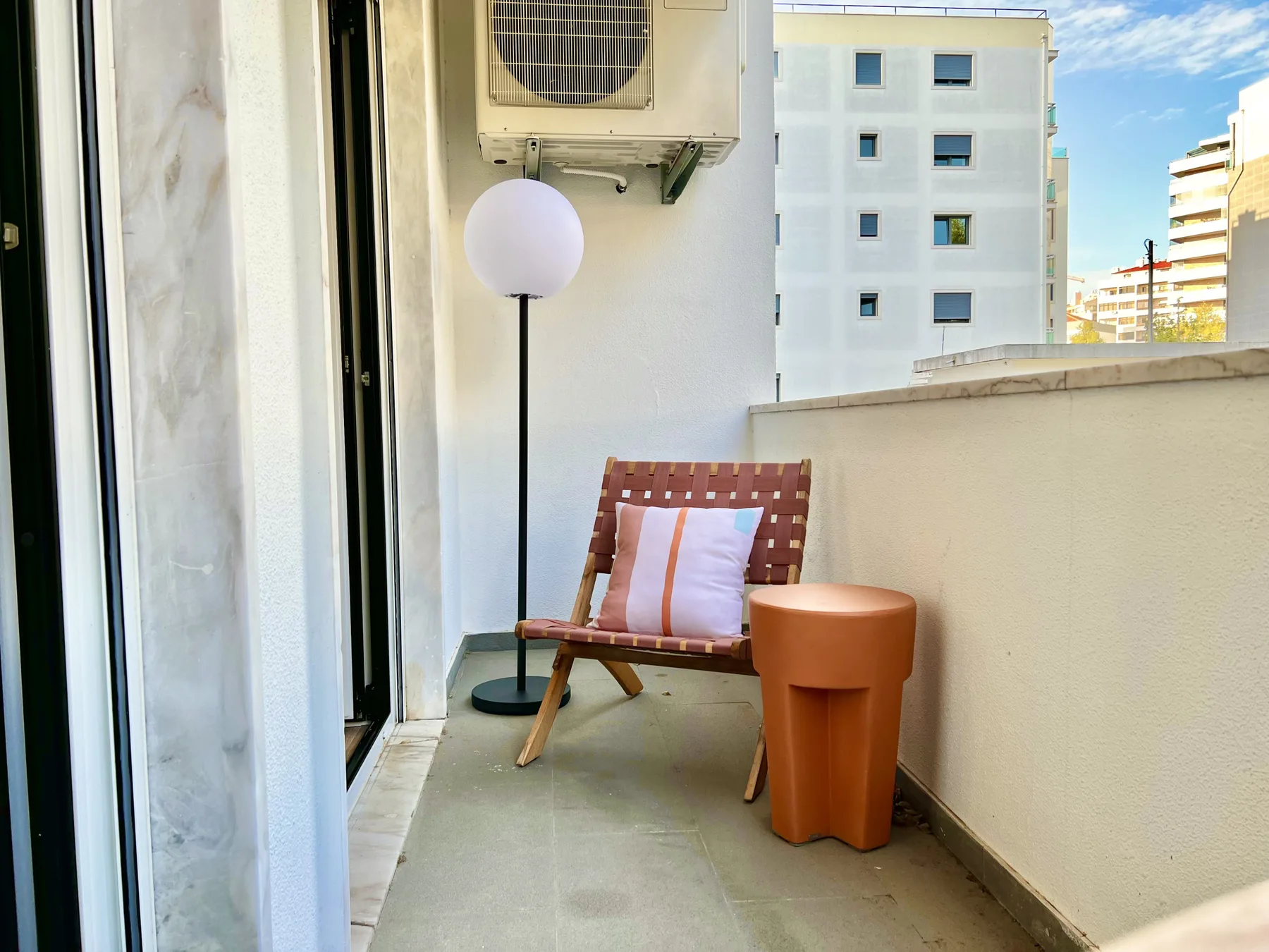 Un balcón estrecho tiene un asiento de madera, una lámpara de pie y una mesita.