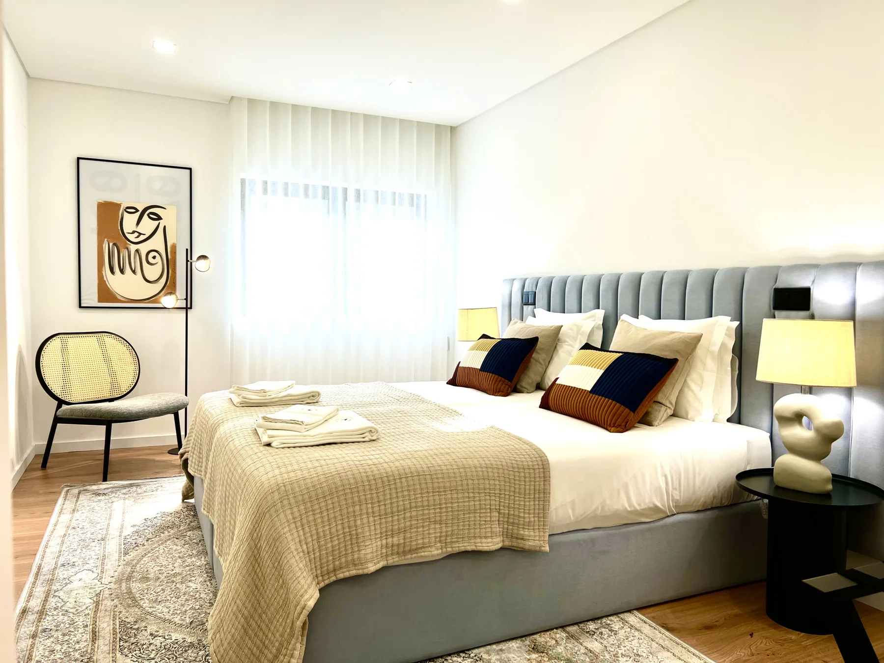 Un dormitorio luminoso con una cama hecha, cojines estampados y una silla junto a la ventana.