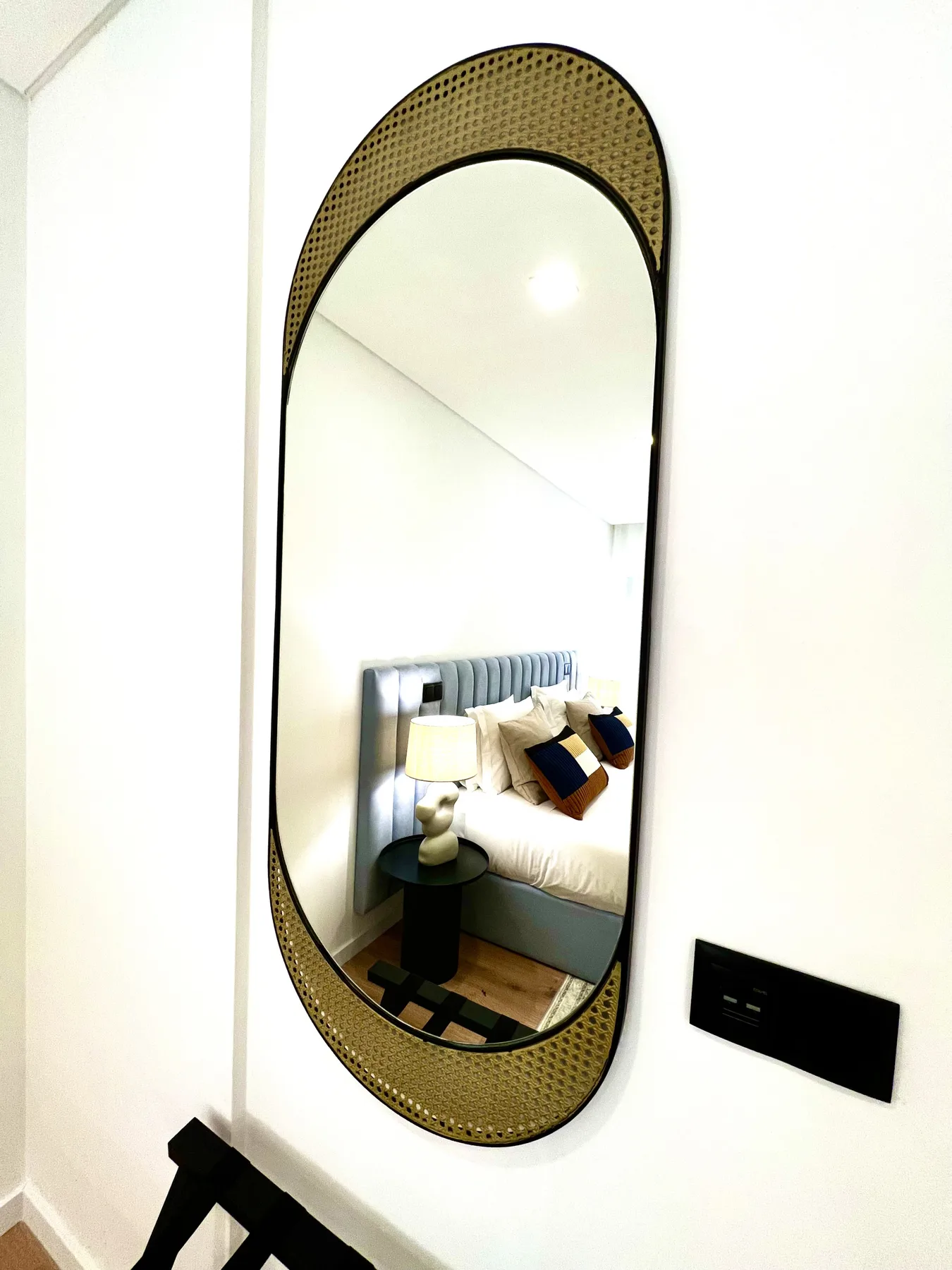Un espejo de pared refleja un dormitorio con una cama y una mesita de noche.
