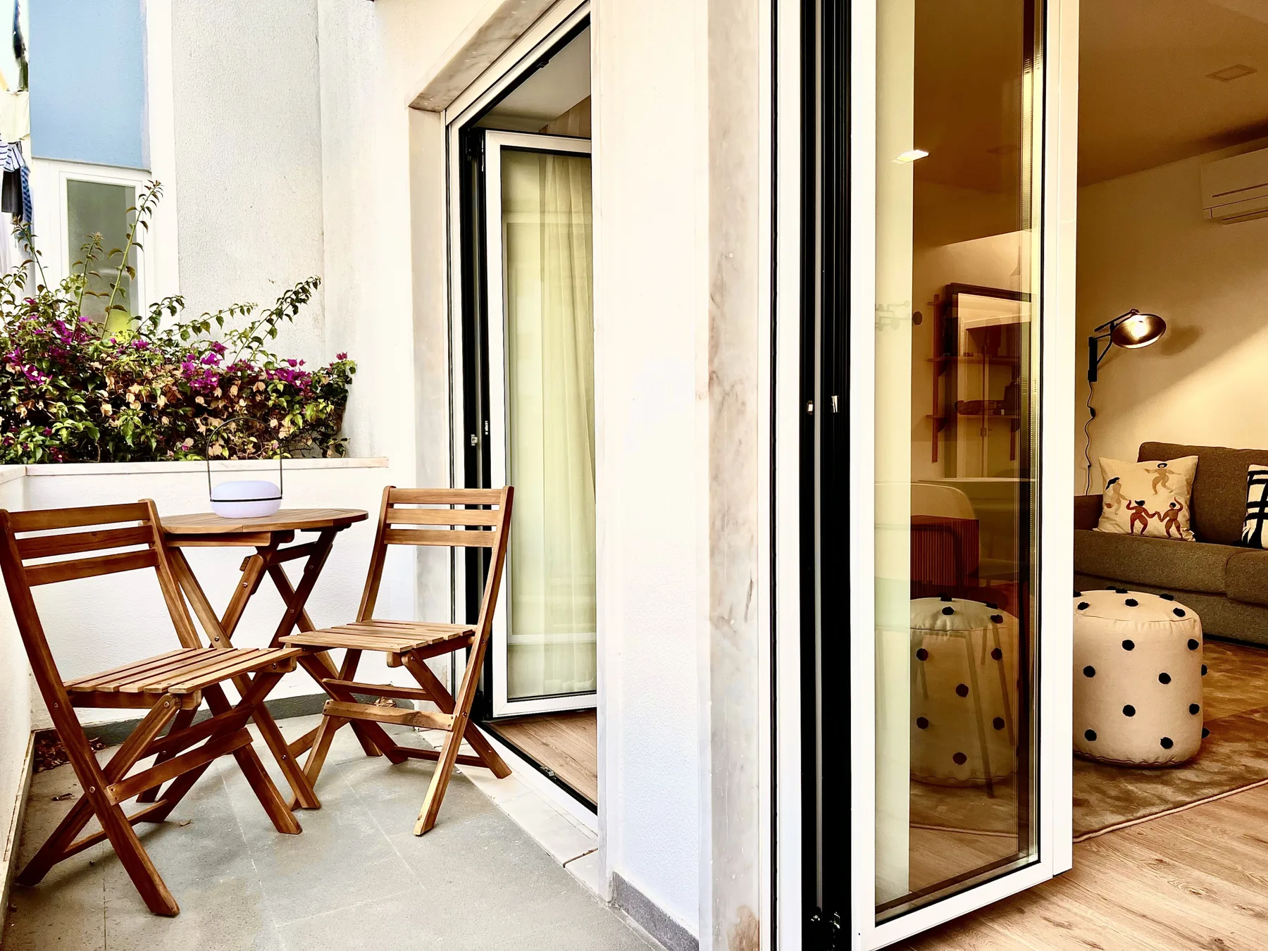 Un pequeño patio con una mesa de madera y asientos junto a una puerta de vidrio abierta.