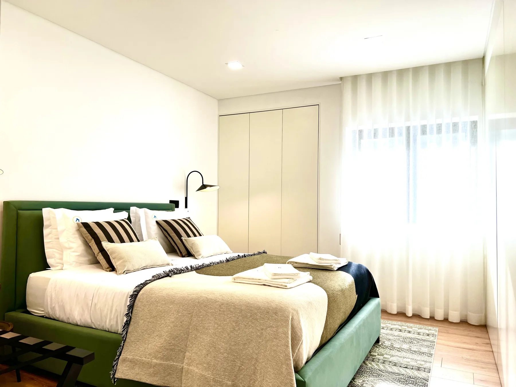 Un dormitorio luminoso con una cama tendida junto a una ventana amplia con cortinas.